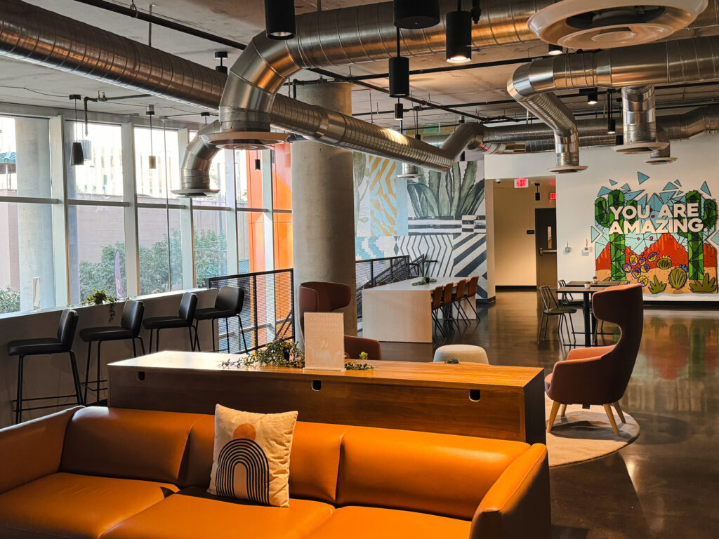 A spacious and modern coworking lounge at Kenect Phoenix featuring sleek industrial design, vibrant wall art with the phrase "You Are Amazing," and a mix of comfortable seating options for socializing or working.