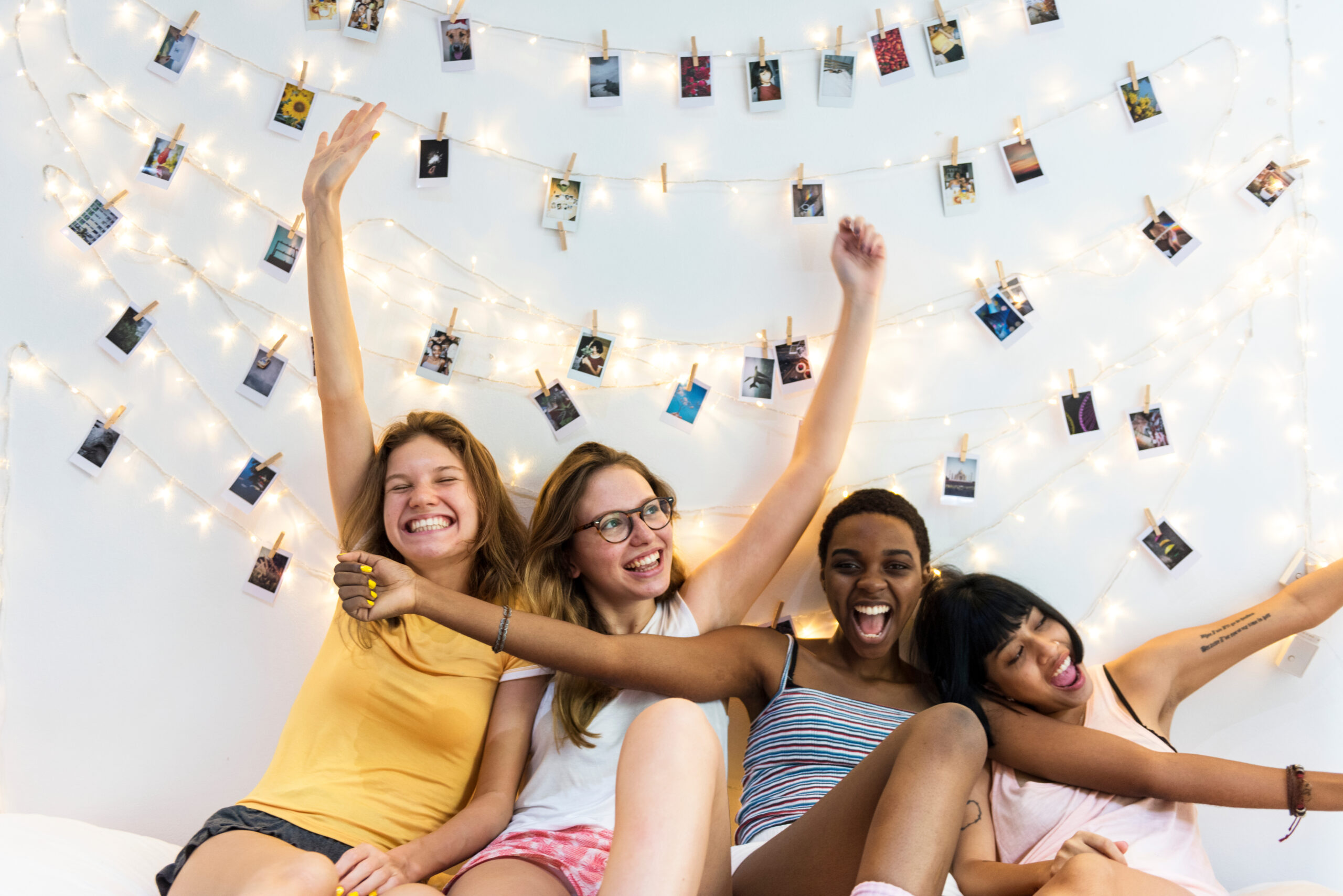 Four female roommates smiling and posing with hands raised in a brightly lit room adorned with string lights and Polaroid photos.