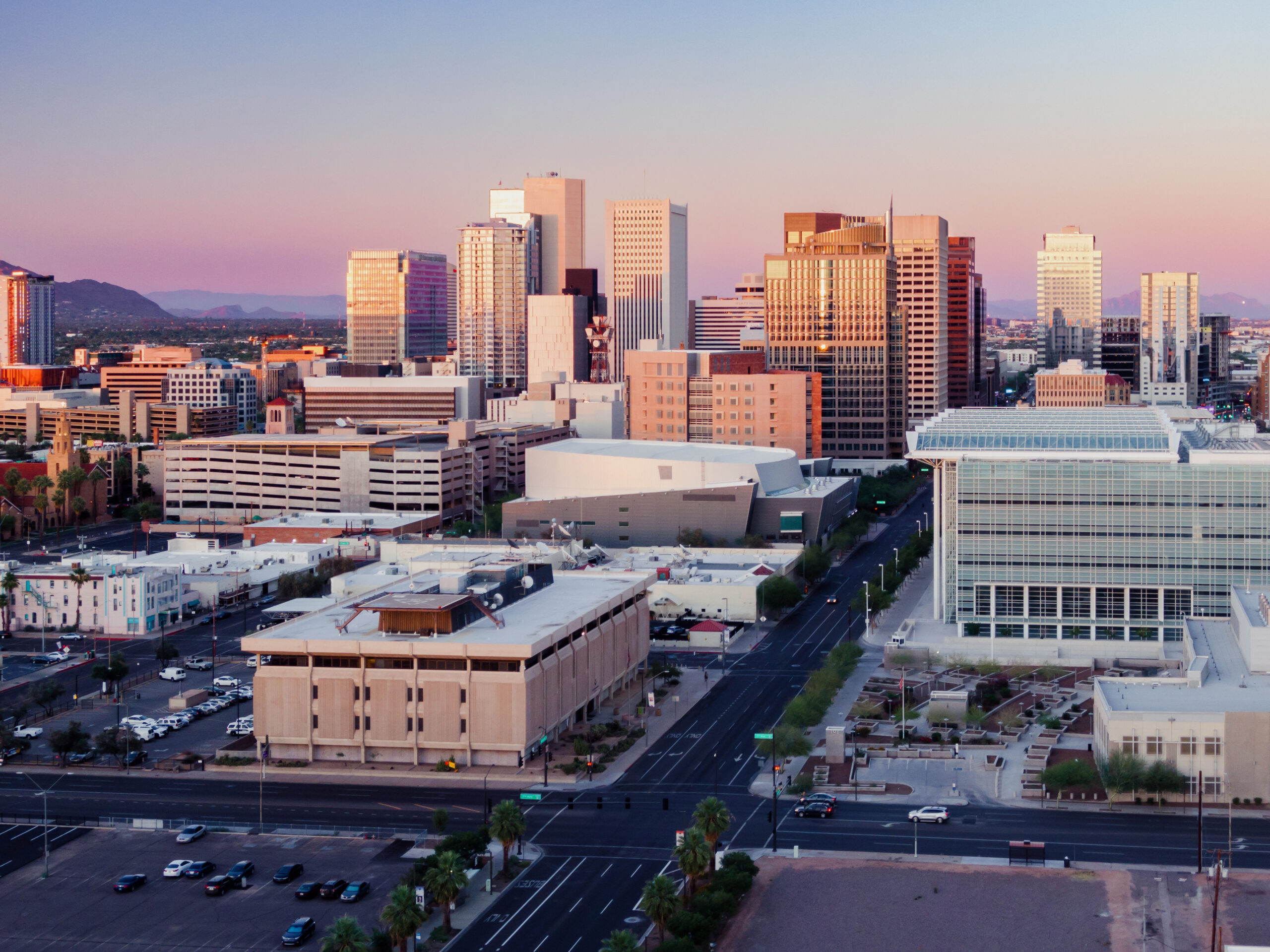  A vibrant cityscape of downtown Phoenix, Arizona, captured at sunset, featuring modern skyscrapers, mid-rise buildings, and distant mountain ranges under a pastel sky.