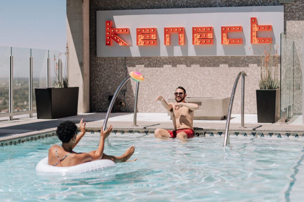 Two male roommates enjoy a sunny day at the rooftop pool at Kenect Phoenix, with one sitting at the pool’s edge and the other floating on an inflatable.