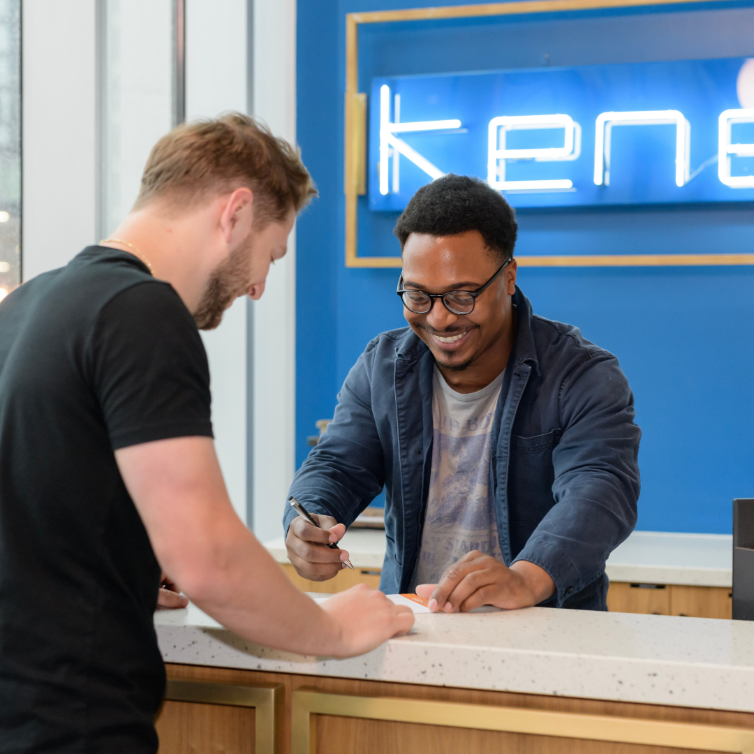 A man with glasses and a friendly smile is helping another man at a counter. The man behind the counter holds a pen, assisting with paperwork.