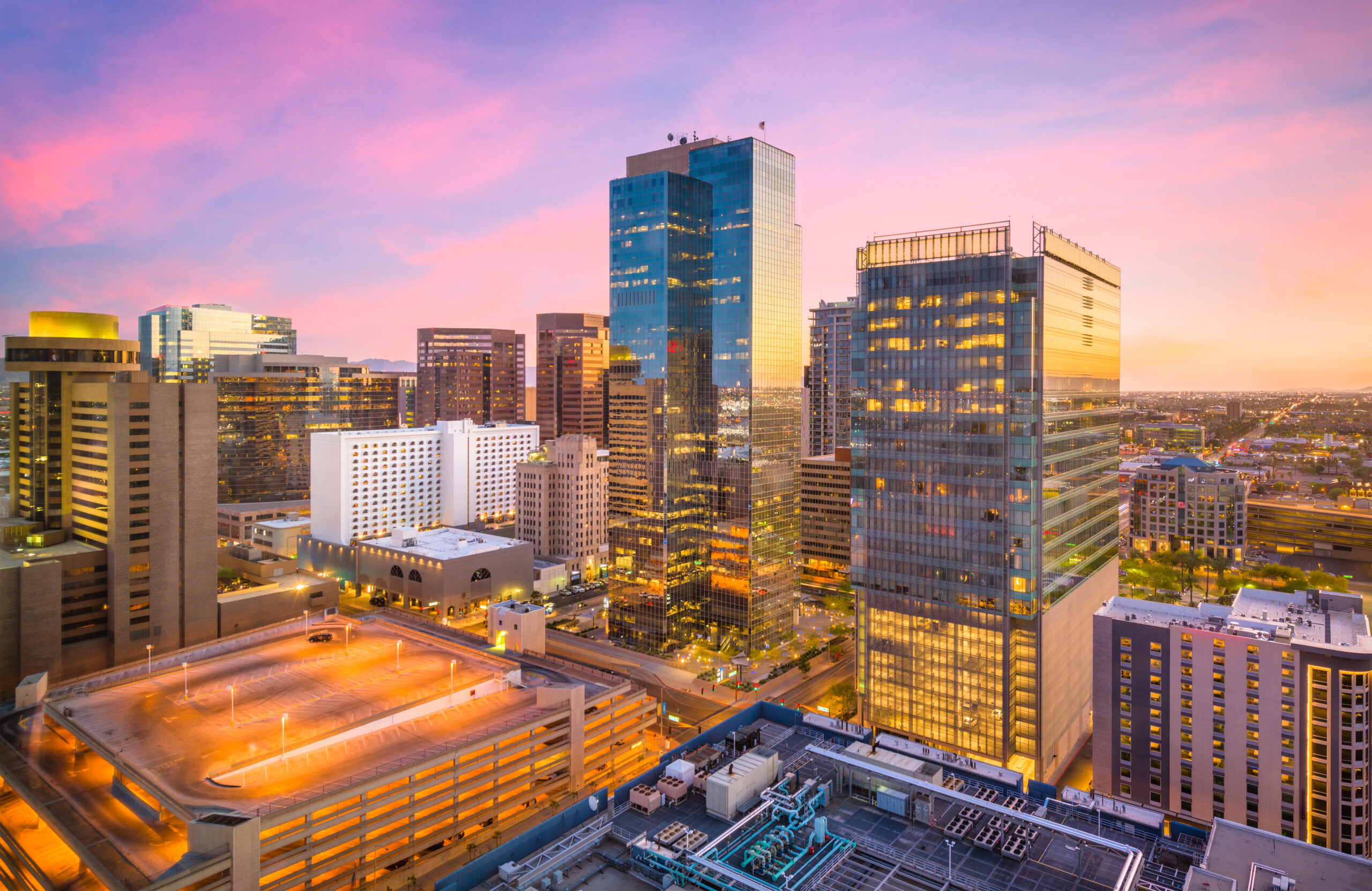 The Phoenix, Arizona skyline at sunset, featuring tall glass skyscrapers reflecting the pink and orange hues of the sky, with a lit parking structure in the foreground.