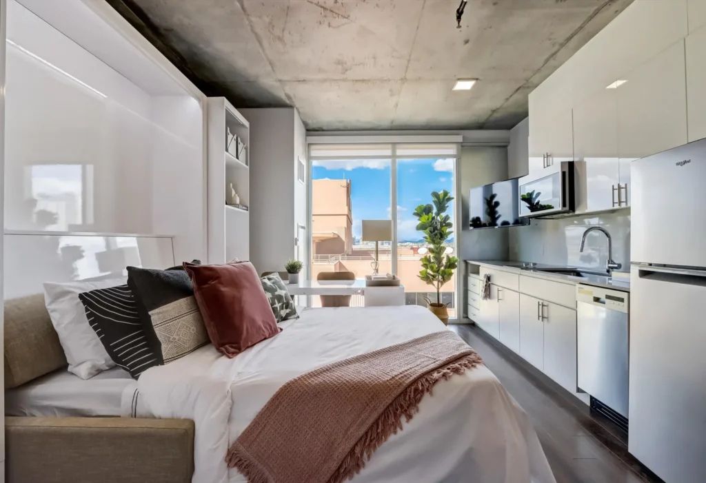 A modern studio apartment at Kenect Phoenix, featuring a neatly made bed with decorative pillows and a throw, a compact kitchen with sleek white cabinets and appliances and a large window offering a view of a sunny cityscape.