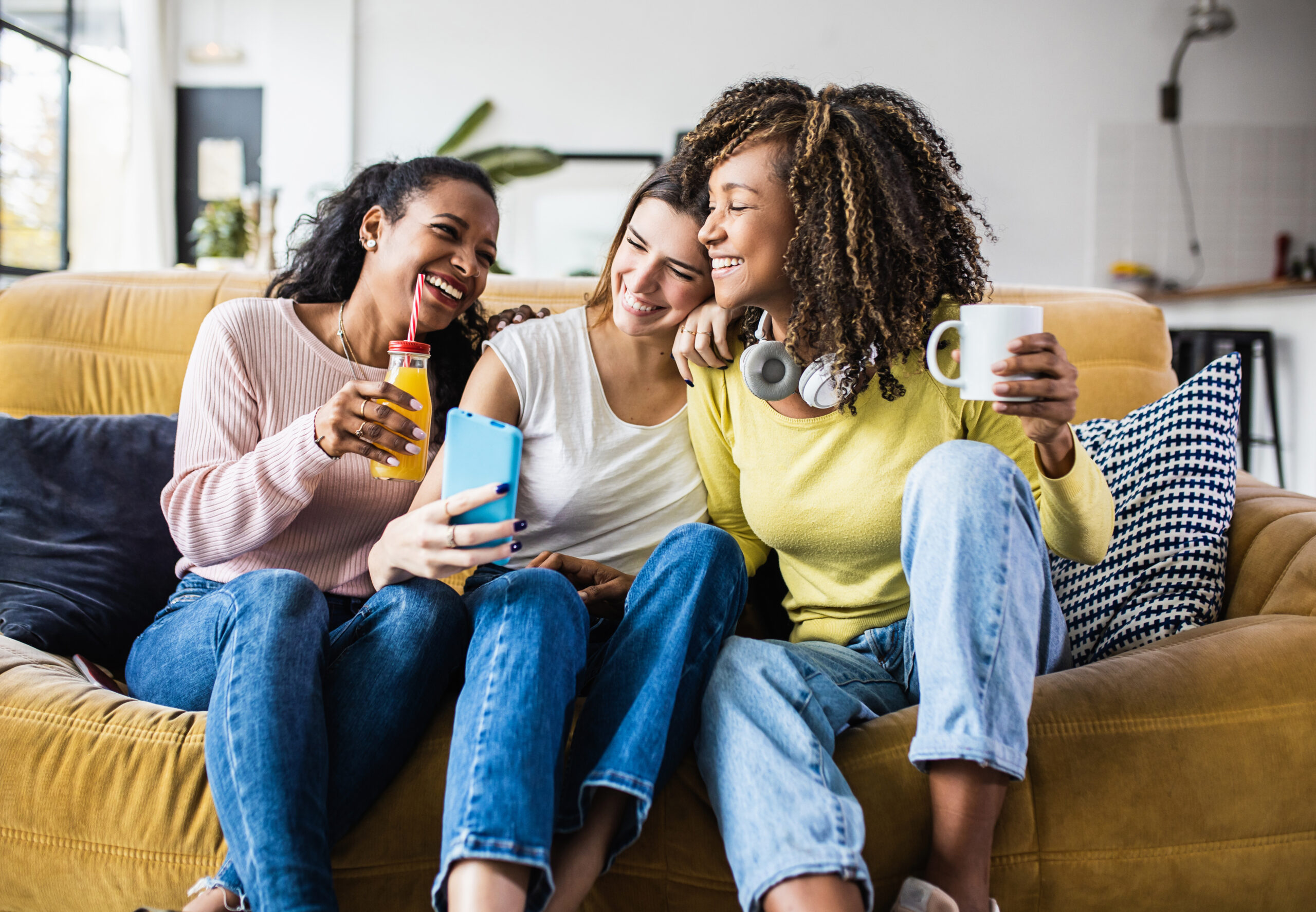 Three friends sitting on a yellow couch, smiling and laughing while taking a selfie with a smartphone. One is holding a bottle of orange juice, another has headphones around her neck and a mug in hand, and the third leans in with a bright smile.