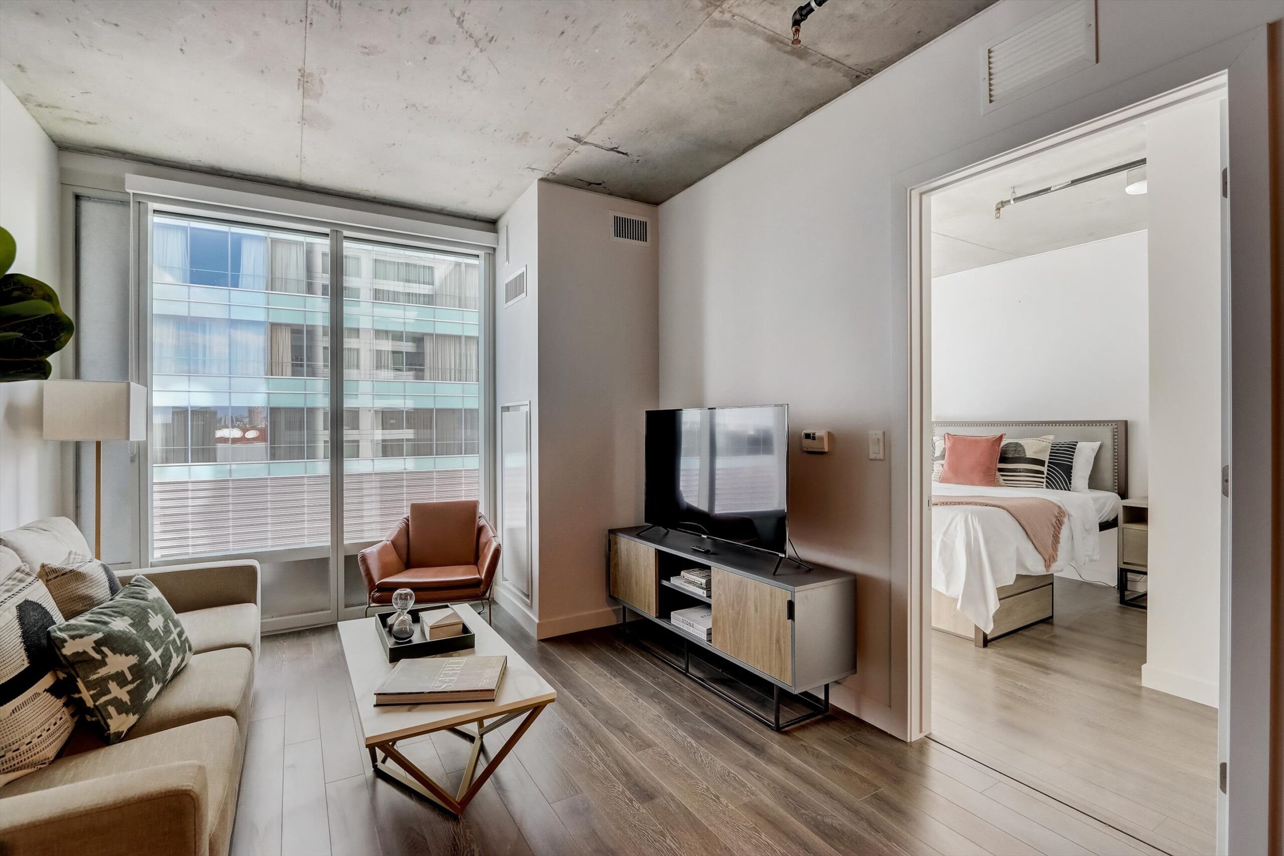 Modern furnished apartment living room at Kenect Nashville, with sofa, coffee table, TV stand, and large windows, leading into a bedroom with a neatly made bed.