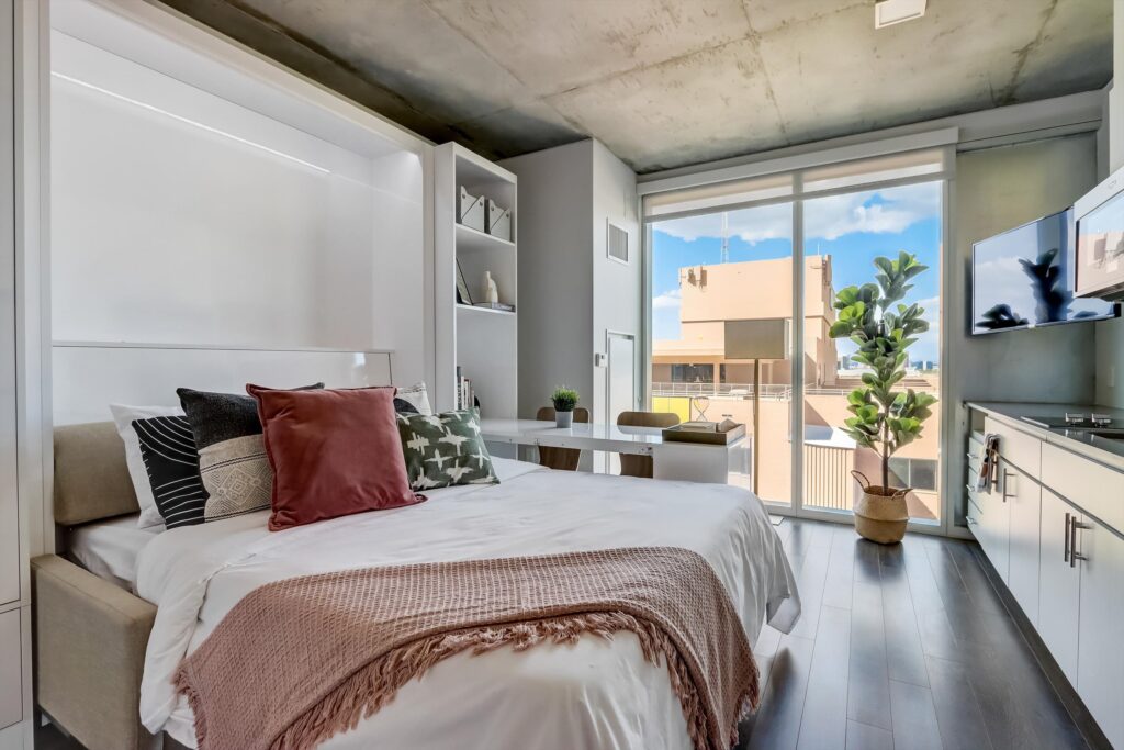 FAQs 1 A bright, modern studio apartment at Kenect with a bed, built-in shelving and expansive floor-to-ceiling windows offering city views.