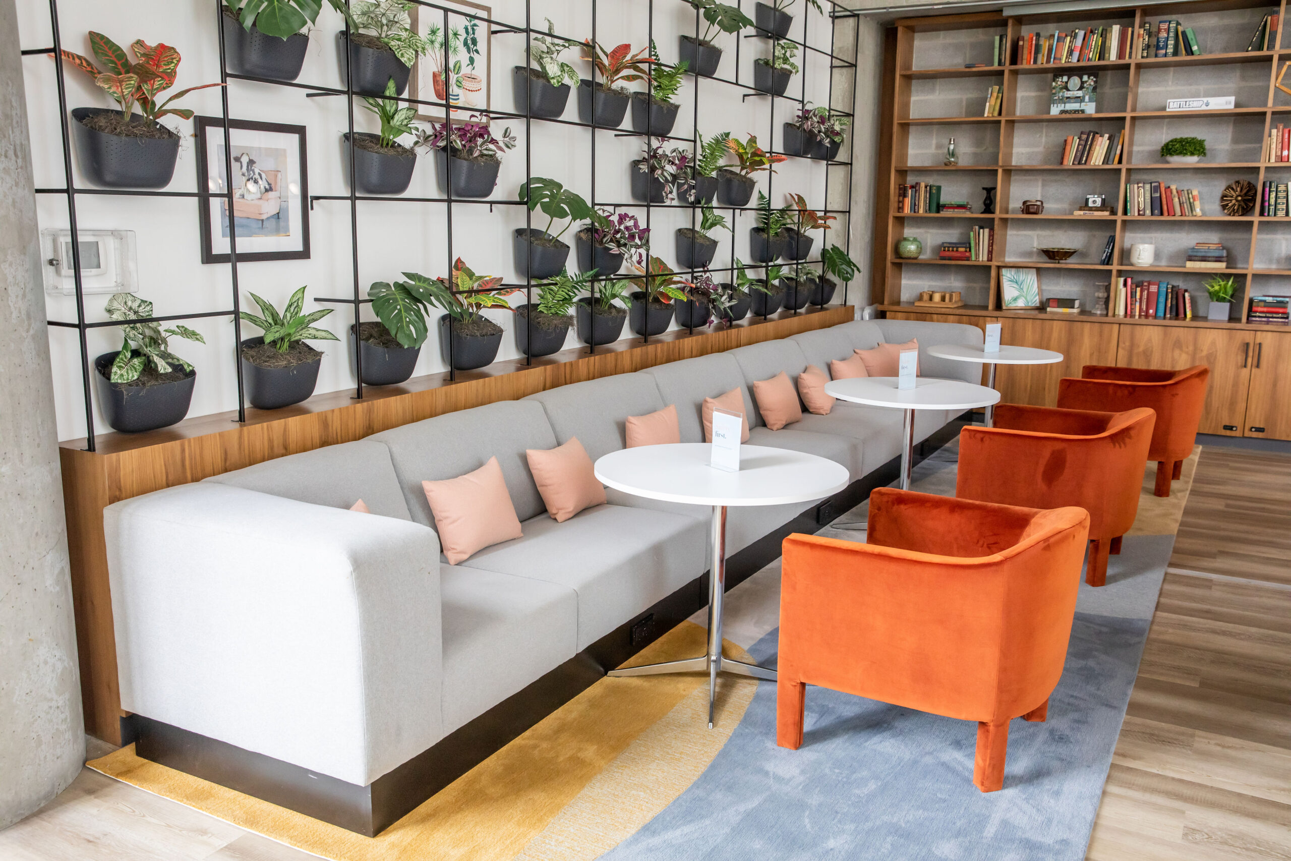From Couch to Cowork: 5 Ways Flexible Coworking Beats Working From Home 2 Bright and modern coworking lounge at Kenect Nashville featuring a long gray sofa with pink pillows, white round tables, orange velvet chairs, a wall of potted plants and built-in bookshelves filled with books and decor.