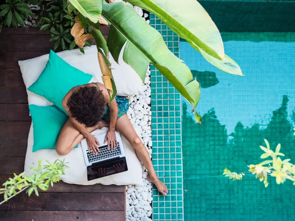 Person working on a laptop while lounging by a turquoise pool, surrounded by plants and outdoor cushions.